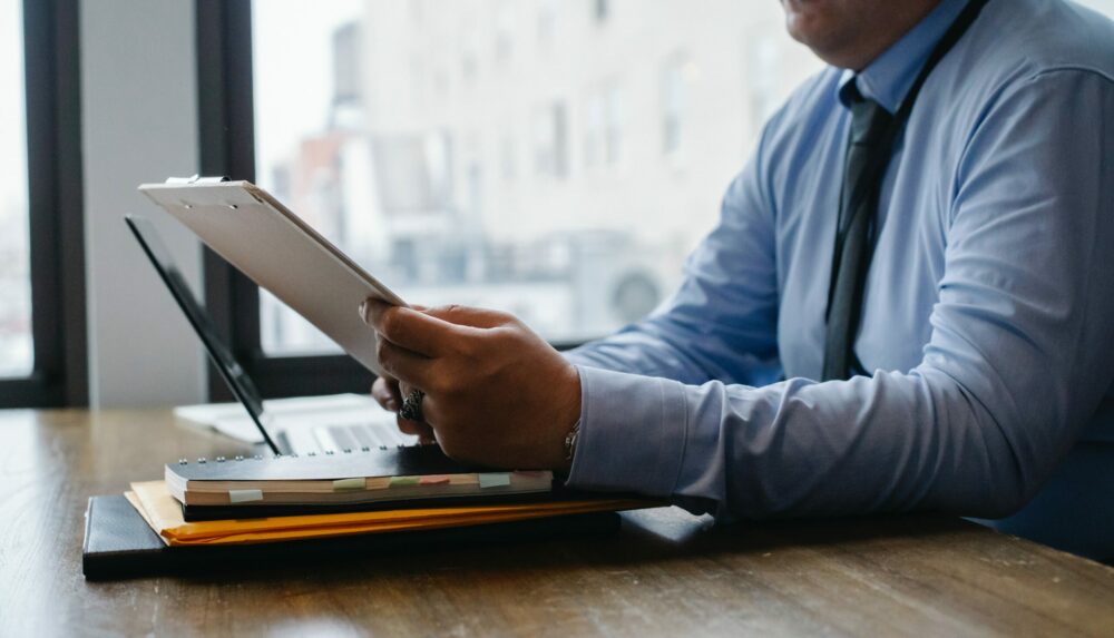 A man in a shirt and tie sitting at a desk with a laptop on it next to a window looking out onto other offices, and holding a tablet.