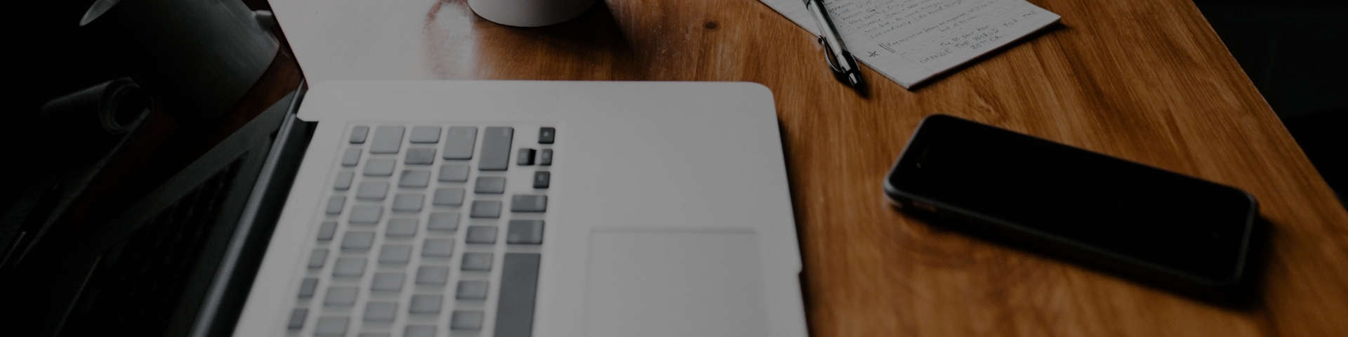 Photo shows a laptop, cup of coffee and notepad and pen on a wooden table by a window
